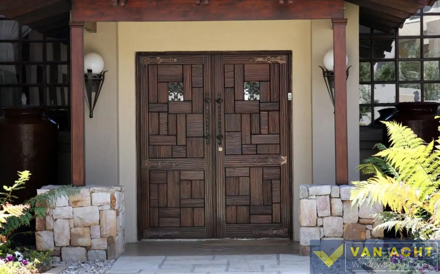 Dark wood double entrance door with a mosaic-style panel pattern and small decorative iron-grille windows, set beneath a covered timber portico with stone-clad pillars.