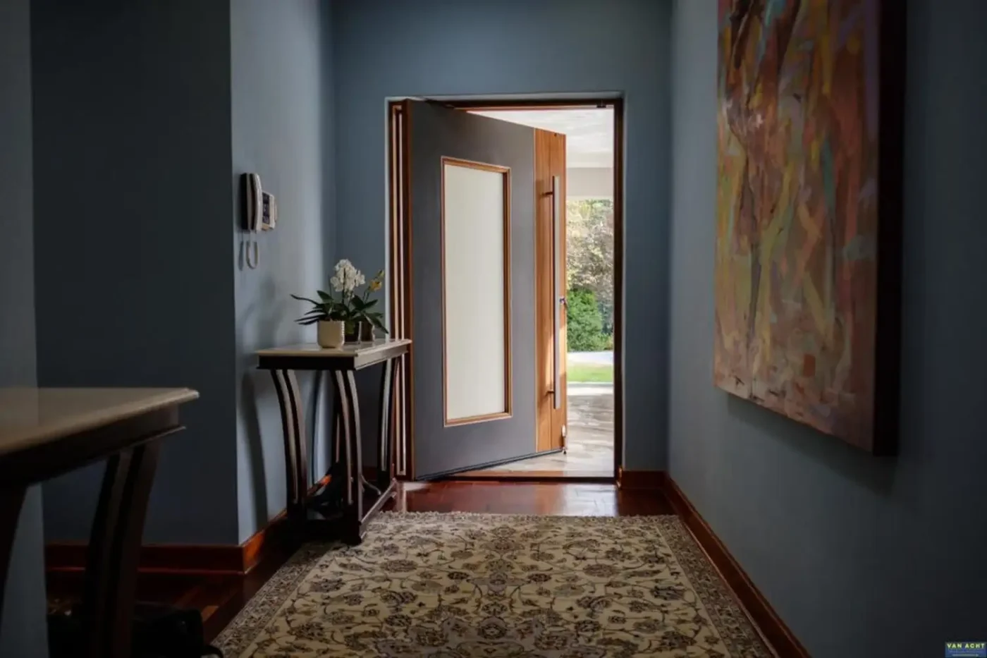 Grey pivot entrance door open onto a hallway, framed in timber with a garden view beyond