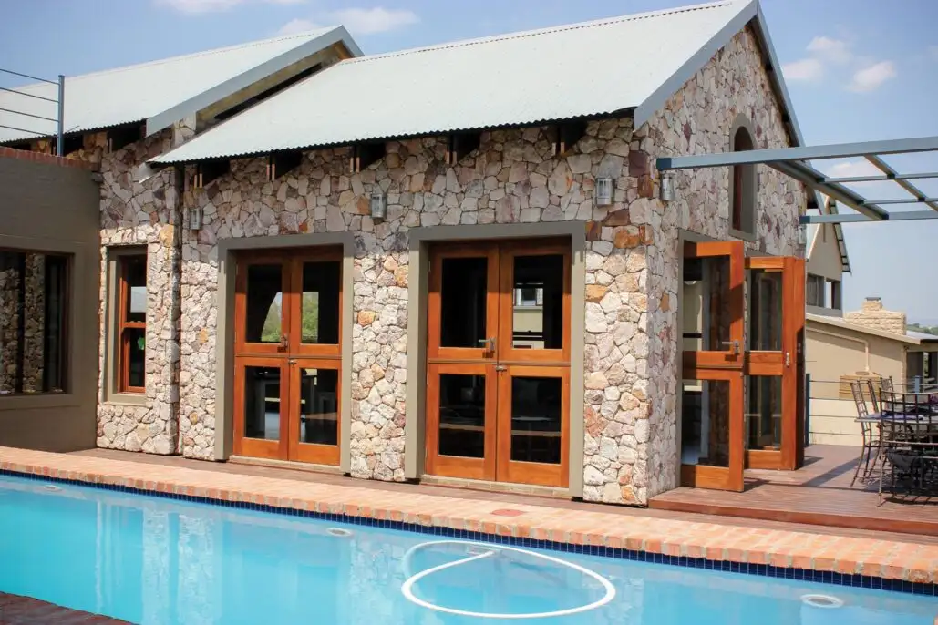 Timber bifold doors and a pivot door on a stone-clad house, opening onto a pool deck.